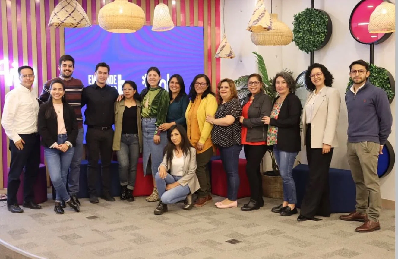 Grupo de participantes y colaboradores del bootcamp con Banco FIE frente a una pantalla de EmprendeBO.